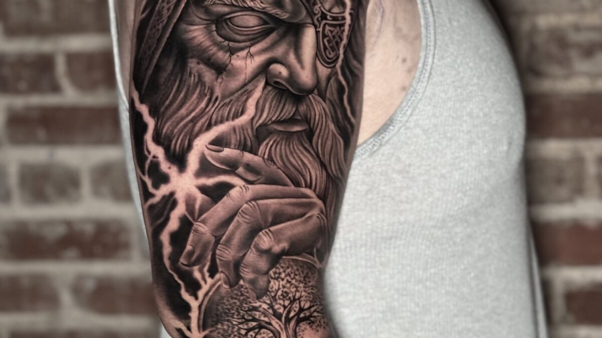 Black and grey Odin realism tattoo by Eike H. Schumacher, showing Odin, a hand, tree imagery, and negative-space lightning across an upper arm.