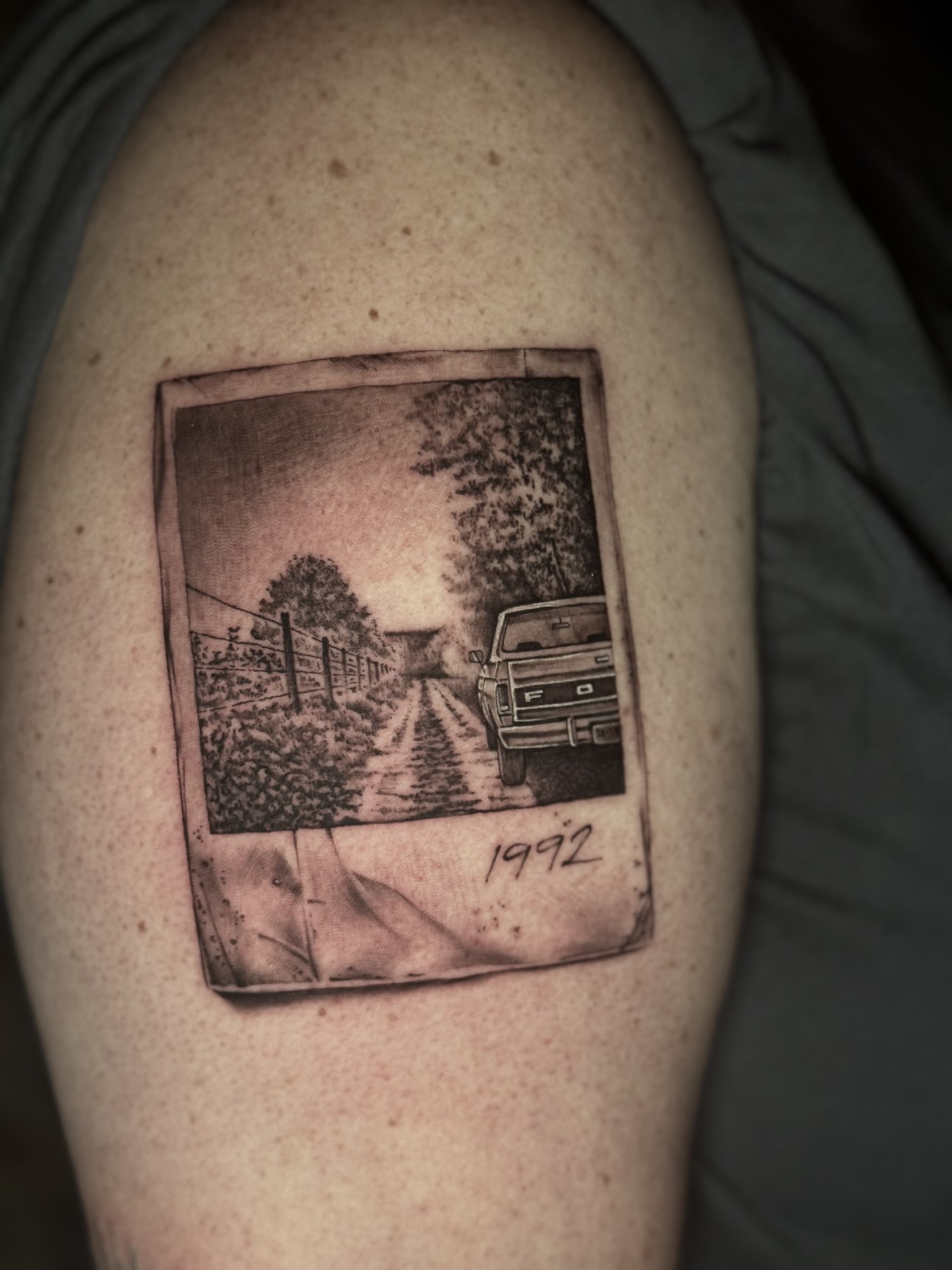 Small black and grey Polaroid realism tattoo by Eike H. Schumacher, depicting a rural road and Ford truck scene.